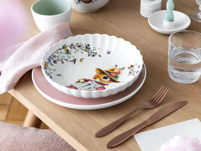 De keramische schaal "Spring Fantasy" met konijnendesign van Villeroy & Boch staat op roze-witte borden, met roségoud bestek en een glas water op een houten tafel.