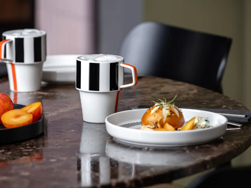 Memphis coffee cups and a Memphis plate with food and a piece of fruit on a marble table next to a black chair.

