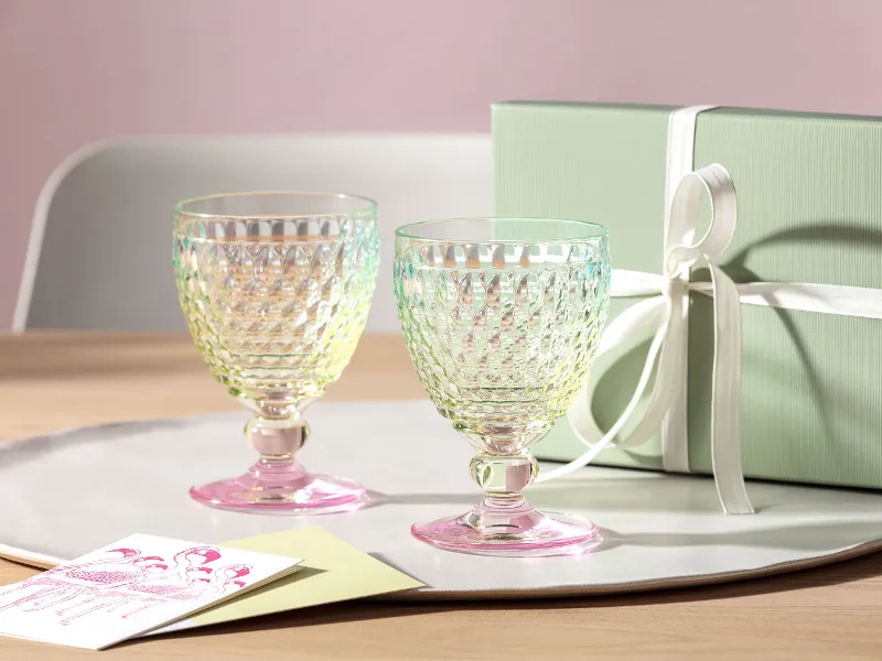 Deux verres Boston de Villeroy & Boch avec un fond rose sont posés sur un plateau à côté d'une boîte cadeau verte avec un ruban blanc et une carte de vœux sur une table en bois.