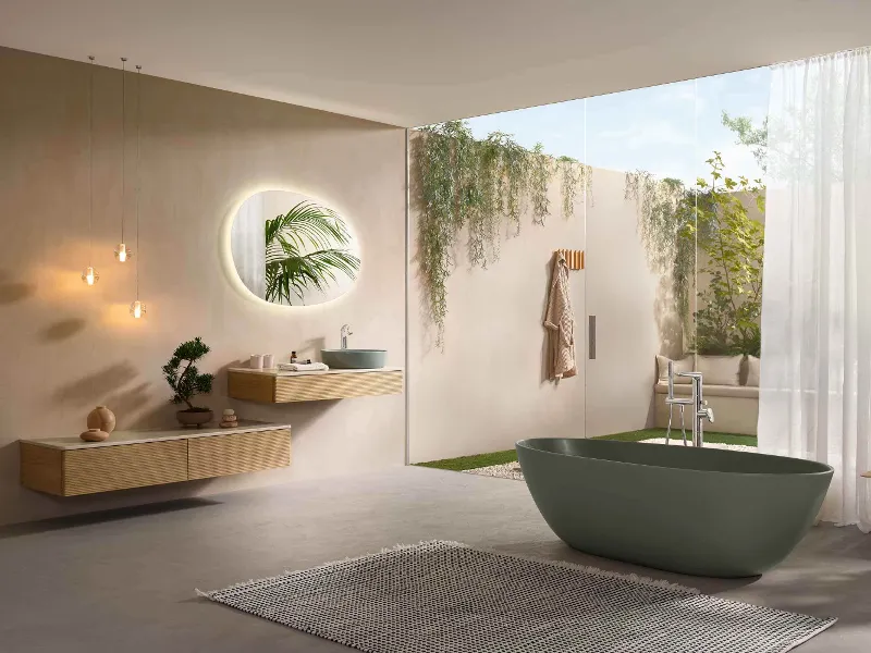 Une salle de bains Antao minimaliste avec robinetterie moderne, un lavabo suspendu en bois, un miroir rond rétroéclairé, des lampes suspendues, une baignoire en îlot, une grande fenêtre, des plantes et une vue sur le jardin.