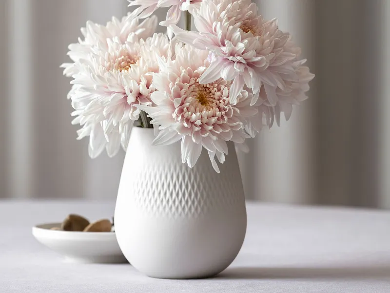 Weiße Manufacture Vase mit blassrosa Chrysanthemen auf einem Tisch, daneben eine kleine Schale mit Steinen.