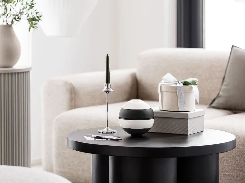 Czarny, okrągły stolik kawowy z La petite Boule marki Villeroy & Boch, zielona świeca na srebrnej podstawce i pudełko upominkowe na dwóch książkach. Beżowa sofa i szary cokół z wazonem w tle.