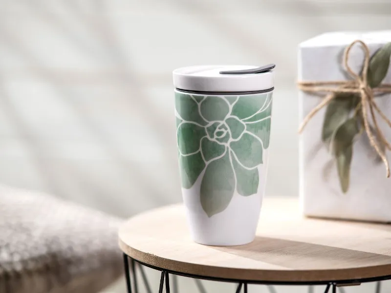 Der Coffee ToGo Becher von Villeroy & Boch, ein Reisebecher aus Keramik mit grünem Blätterdesign, steht auf einem Holztisch neben einer mit Bindfaden zusammengebundenen Geschenkbox.