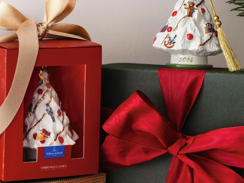 Christmas decoration from Villeroy & Boch with a decorated tree on a base with the inscription “2024”, presented together with a red box and a wrapped gift with a large red bow.