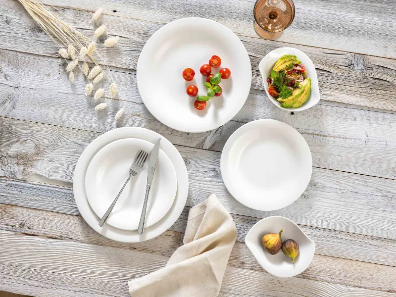 A rustic table setting with Essential White and New Cottage white plates, a fork and a knife, a beige napkin and bowls of cherry tomatoes, avocado slices and figs. On the top right there is a glass of rosé wine.