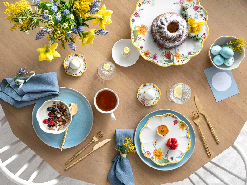 Ein frühlingshaft gedeckter Tisch mit Spring Awakening und Crafted Geschirr, einem Gugelhupf, einer Schüssel Haferflocken mit Beeren, einer Tasse Tee und dekorativen Eiern.