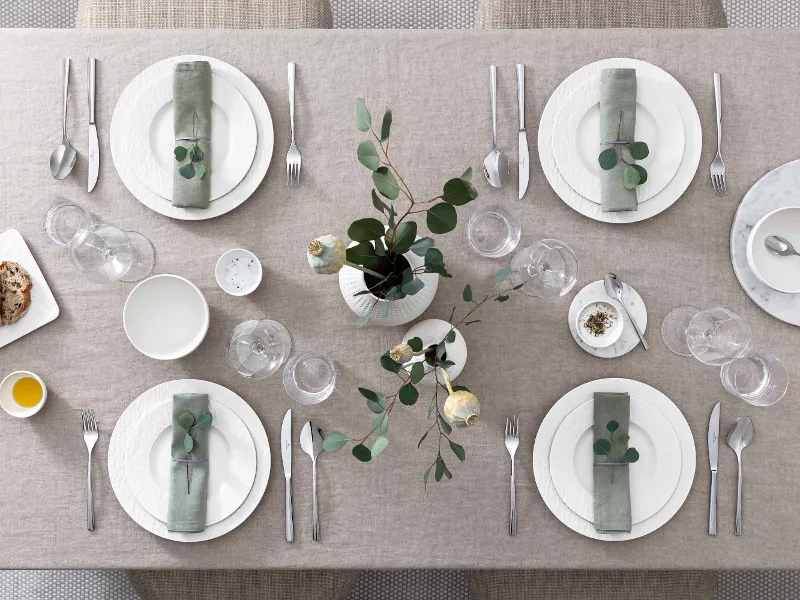 Tavolo da pranzo apparecchiato con eleganza con stoviglie e bicchieri della collezione Manufacture in bianco e un semplice centrotavola verde e bianco.