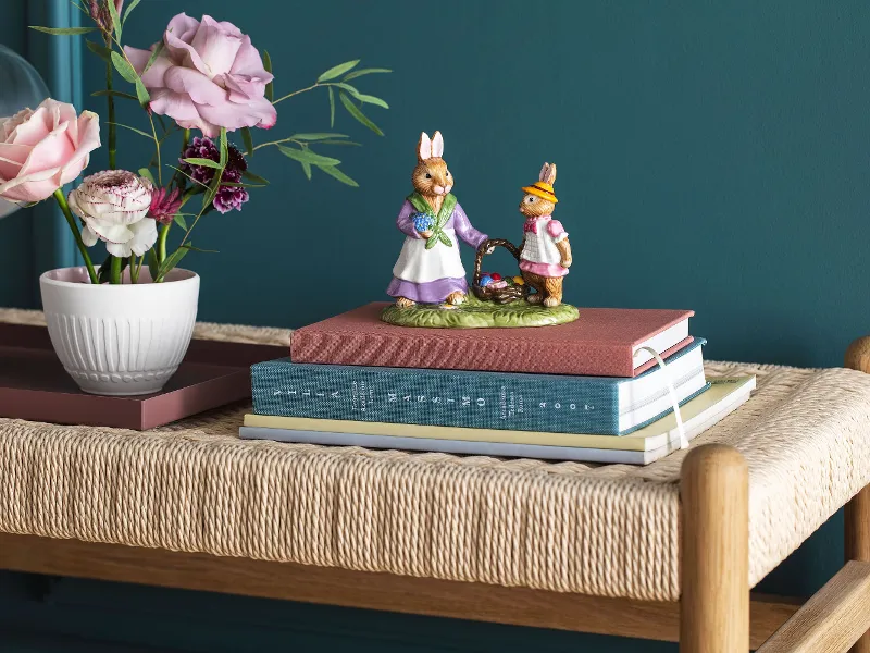 De Spring Fantasy-collectie van Villeroy & Boch toont een konijnenfiguur op boeken naast een vaas met roze en paarse bloemen, geplaatst op een gevlochten bank voor een blauwgroene muur.