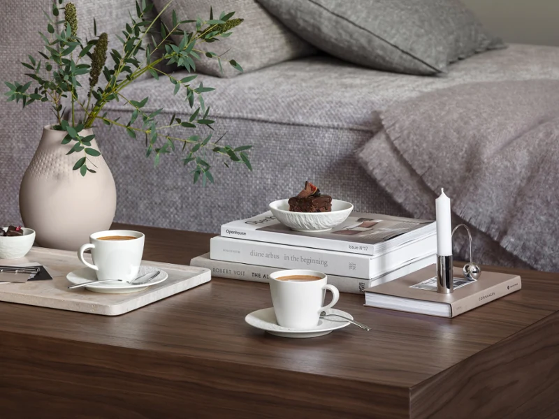 A cozy living room with a wooden coffee table, on which stand two Manufacture coffee cups, a small dessert, a stack of books, a candle and a Manufacture Collier vase with green plants.