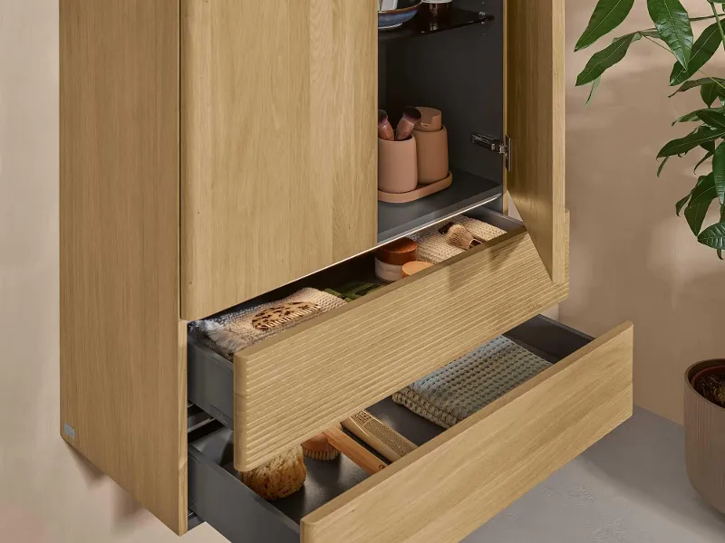 Armadietto per il bagno in legno chiaro con due ante e due cassetti aperti di Villeroy & Boch su una parete naturale.