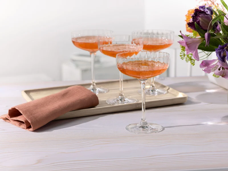 Quatre verres coupe Rose Garden avec des cocktails orange sur un plateau. Quatre verres coupe Rose Garden avec des cocktails orange sur un plateau, à côté d'une serviette pliée et d'un vase de fleurs sur une table blanche.