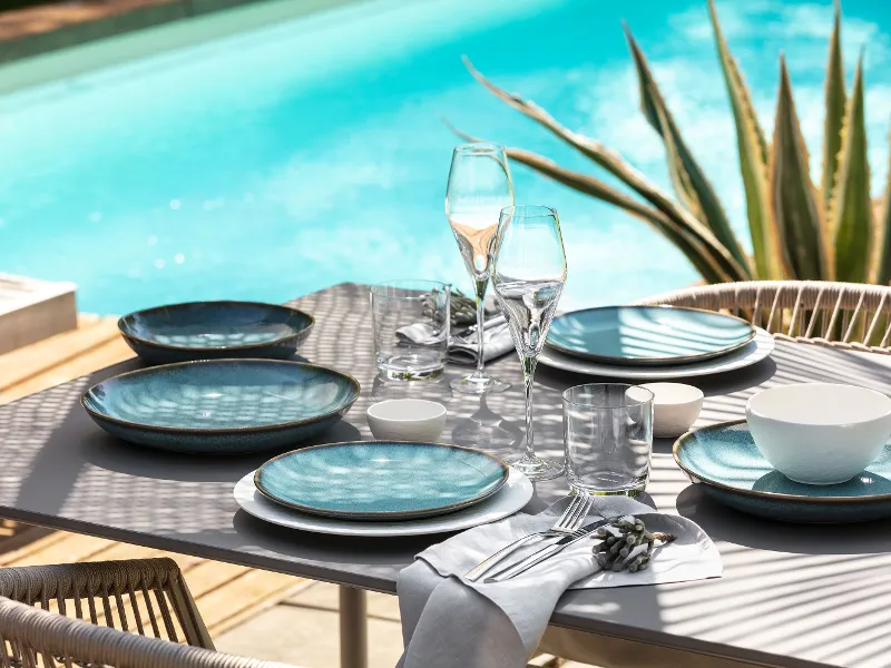 Das Tischset „Lave“ von Villeroy & Boch mit blauen Tellern, Gläsern und Besteck blickt auf einen türkisfarbenen Pool, während Pflanzenschatten Muster auf der Oberfläche erzeugen.	