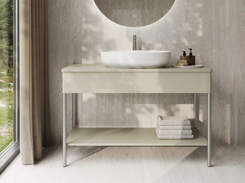 Baño moderno con un espejo redondo, un lavabo sobre encimera, un mueble de lavabo blanco y toallas dobladas, situado junto a una gran ventana con cortinas.