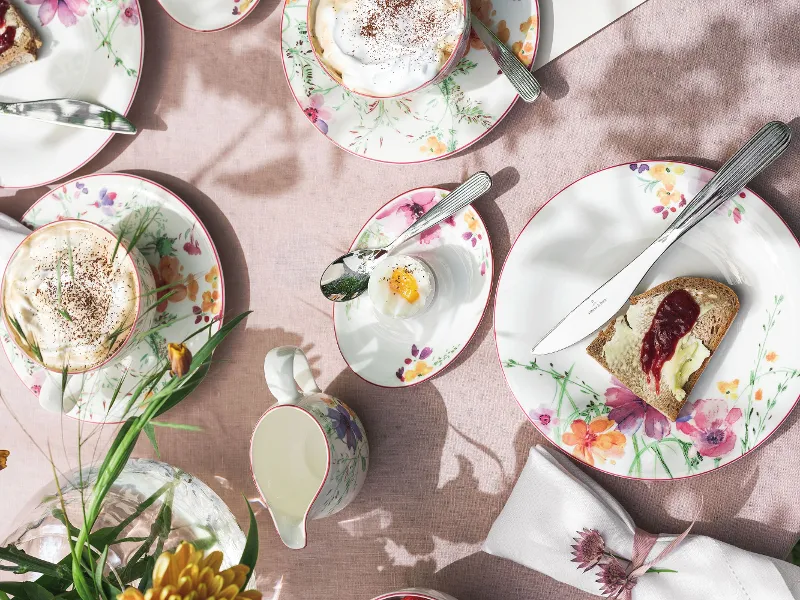 Un set da tavola Mariefleur di Villeroy & Boch con pane tostato e marmellata, un uovo in una tazza e cappuccini. Una lattiera e fiori decorano la tovaglia rosa.