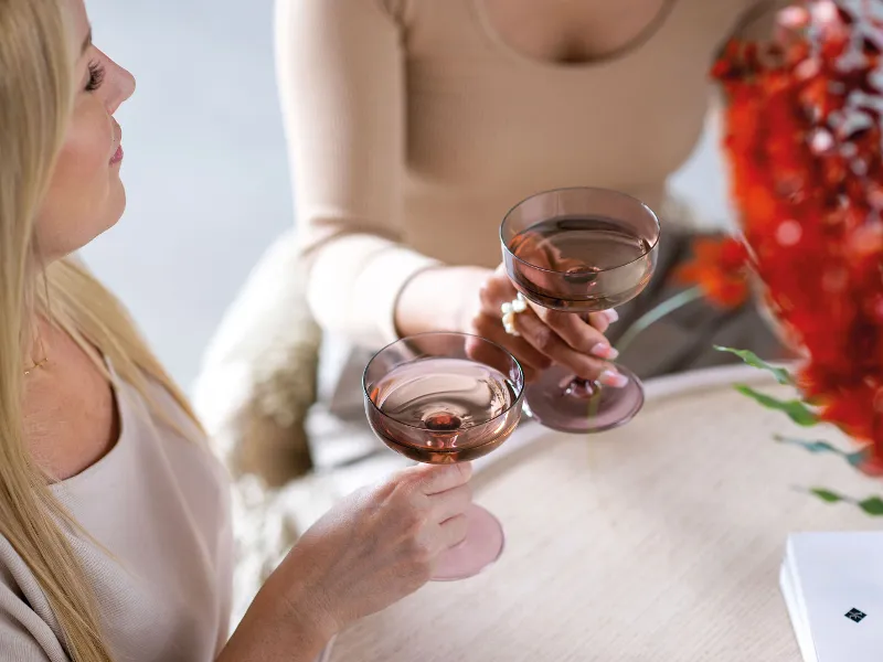 Two people holding Like Glass glasses filled with drinks from Villeroy & Boch and sitting at a table decorated with a red flower arrangement and a book with the inscription ‘Mallorca’.	