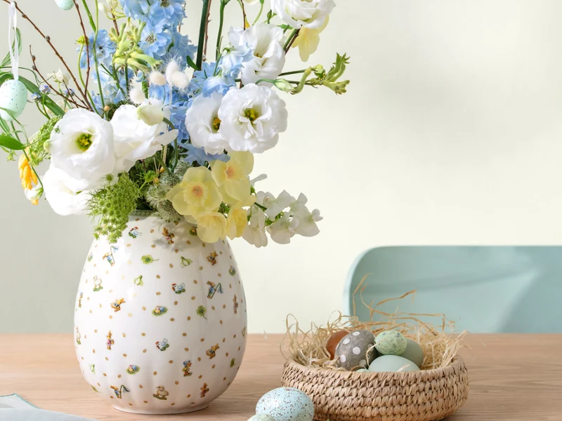 Un vase Easter Delight avec des fleurs blanches et bleues est posé à côté d'un petit nid rempli d'œufs pastel sur une table en bois.