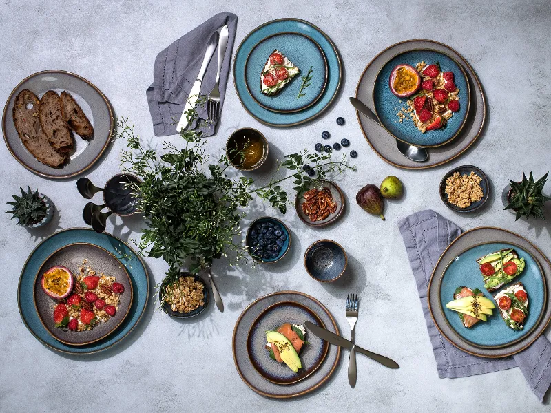 Bovenaanzicht van een goed gedekte eettafel met Lave borden vol gezonde maaltijden, fruit en decoratieve planten op een gestructureerd oppervlak.