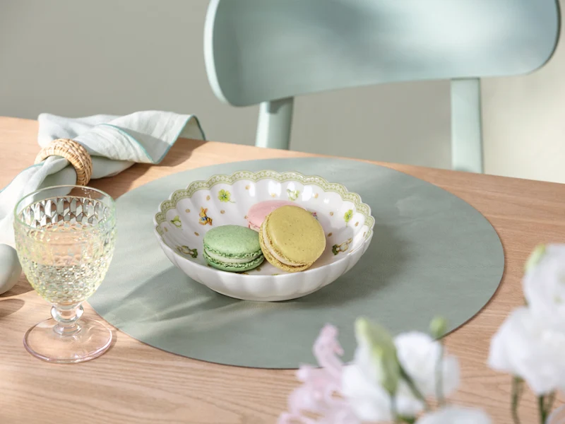 Une coupe Easter Delight remplie de macarons pastel repose sur un set de table Easter Delight vert clair, à côté d'un verre et d'une serviette pliée, sur une table en bois