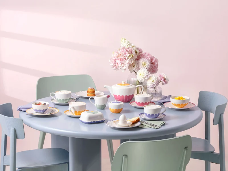 Tavolo rotondo blu pastello con sedie color pastello, apparecchiato per il tè, con tazze e piatti Fleur con motivi floreali, pasticcini e un vaso con fiori rosa al centro.