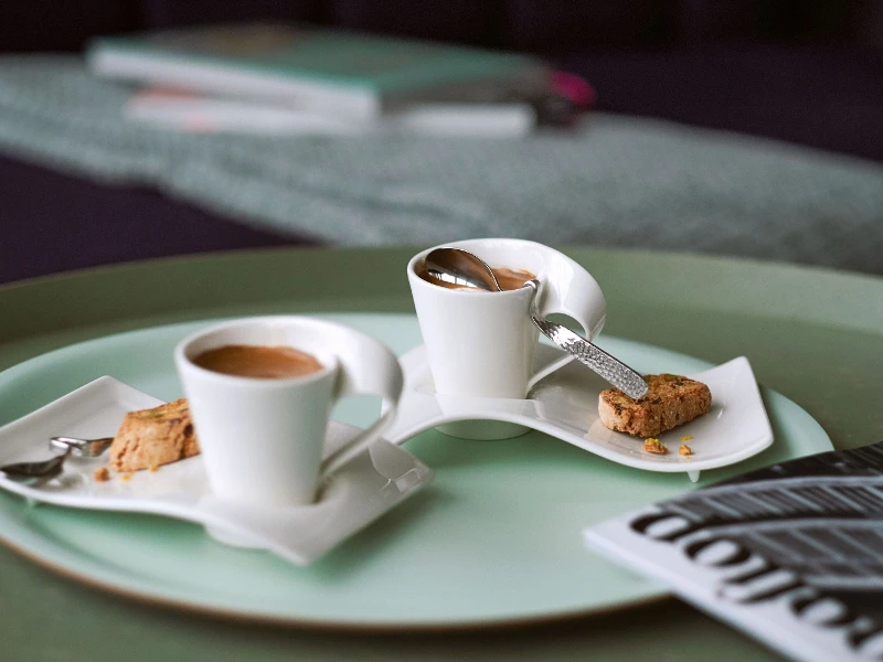 Zwei NewWave Espressotassen mit Löffel und Unterteller, auf einem großen grünen Tablett mit einer Zeitung daneben.