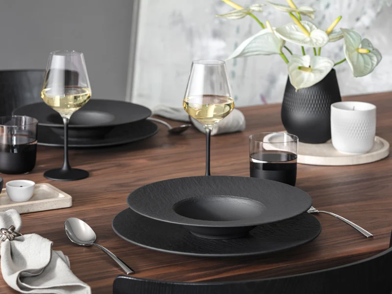 A modern wooden dining table set with Manufacture tableware
A modern wooden dining table set with Manufacture tableware, wine glasses, cutlery, and a vase with white flowers.
