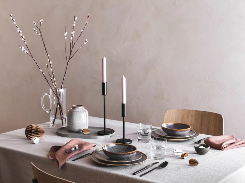 Une table de salle à manger élégante avec des bougies allumées, de la vaisselle en céramique Lave, des couverts de table Manufacture noirs et un décor de Pâques minimaliste avec des chatons de saule.