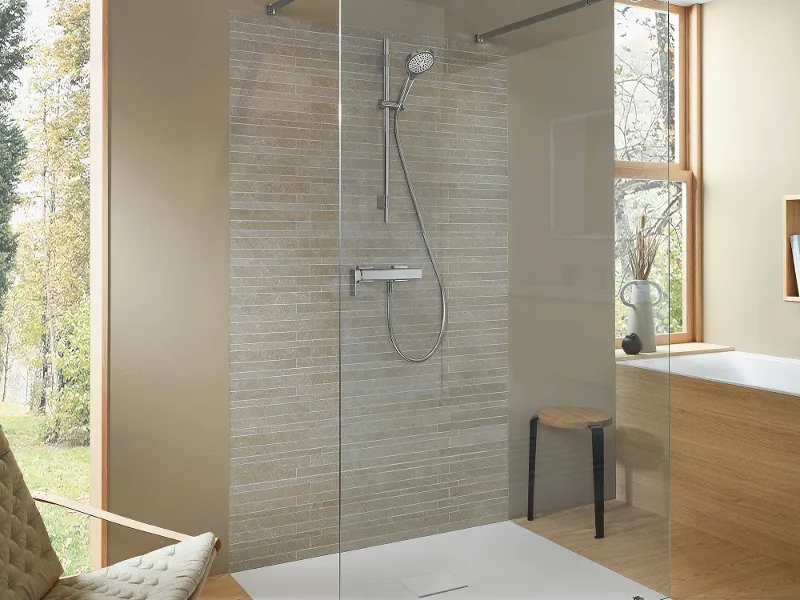 Modernes Badezimmer mit verglaster Dusche mit Handbrause, beige gefliester Akzentwand, hellem Holzboden und einem Stuhl. Große Fenster und Einbauleuchten sorgen für natürliches Licht.