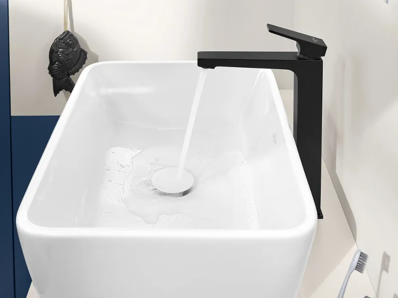 Une salle de bains avec un lavabo rectangulaire blanc Architectura, de l’eau s’écoule du robinet noir. À droite, se trouvent un gobelet bleu avec une brosse à dents et un tube de dentifrice, à gauche, en arrière-plan, une petite serviette sombre est suspendue à un crochet.