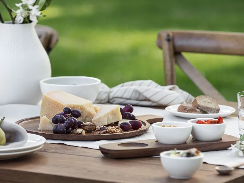 Un élégant couvert Artesano de Villeroy & Boch se compose d'une table en bois décorée de fromage, de raisins, de noix et de pain. Des coupes en céramique stylées et un vase blanc avec des fleurs rehaussent le décor rustique.