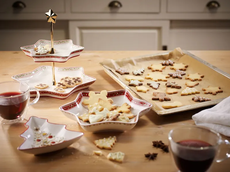 Ein Holztisch mit sternförmigen Schalen von Villeroy & Boch Winter Bakery Delight, auf denen dekorierte Kekse liegen, und einem Backblech mit festlichen Keksen auf Backpapier. Auf dem Tisch stehen zwei Gläser mit rotem Getränk.