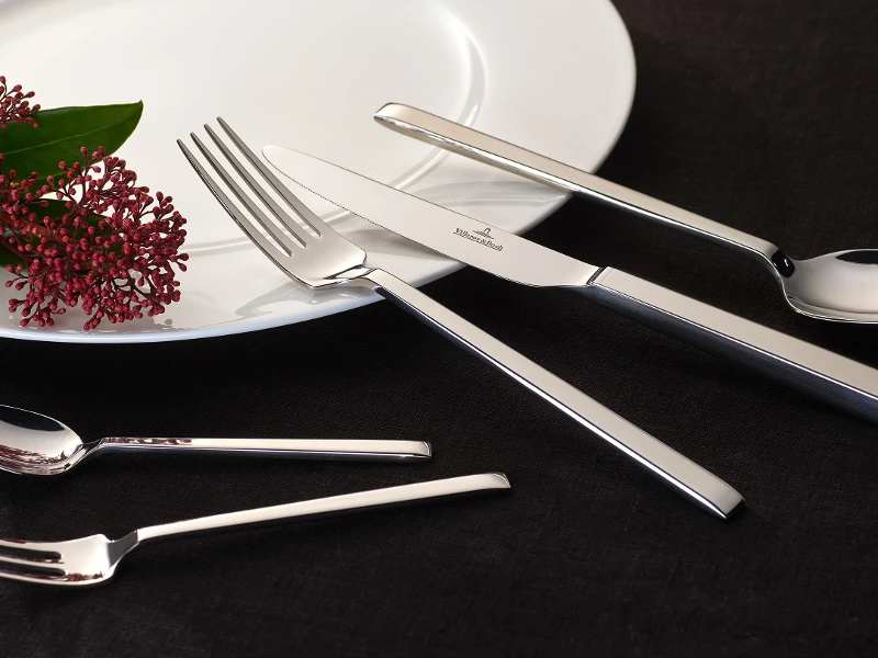 Des couverts de table La Classica brillants et une fleur décorative sur une assiette blanche devant un fond sombre.