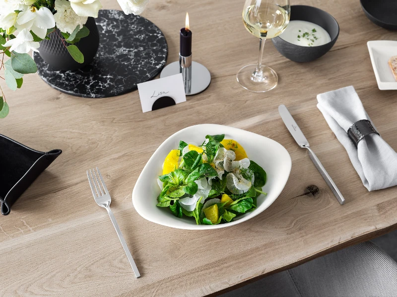 A Vapiano bowl from Villeroy & Boch with salad on a wooden table, next to it a knife and fork from NewWave.