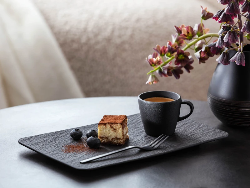 Una fetta di tiramisù accanto a una tazzina da espresso nera Manufacture, guarnita con mirtilli su un moderno piatto rettangolare Manufacture con un vaso decorativo e fiori sullo sfondo.