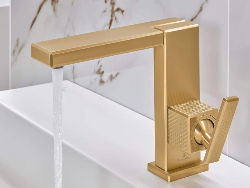 A modern golden faucet in the bathroom with running water in front of a white and marble backsplash.