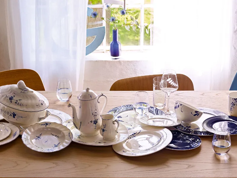 Une table en bois dressée avec une vaisselle en porcelaine bleue et blanche assortie, des tasses et une théière. Des verres d’eau sont disposés parmi la vaisselle. Une fenêtre avec des rideaux légers se trouve en arrière-plan.
