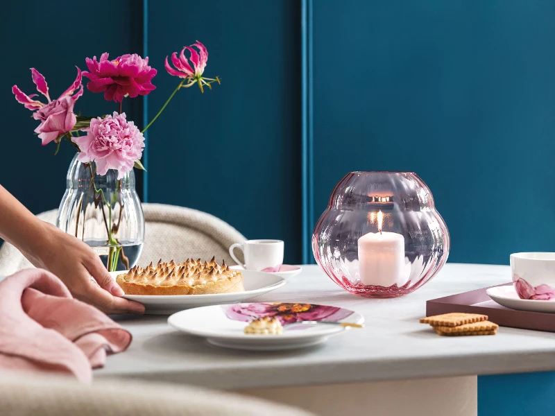 Eleganter Esstisch mit einer Person, die das Dessert serviert, frischen Blumen in Rose Garden Vasen und Tellern aus der Kollektion Rose Garden, die das Ambiente verstärken.