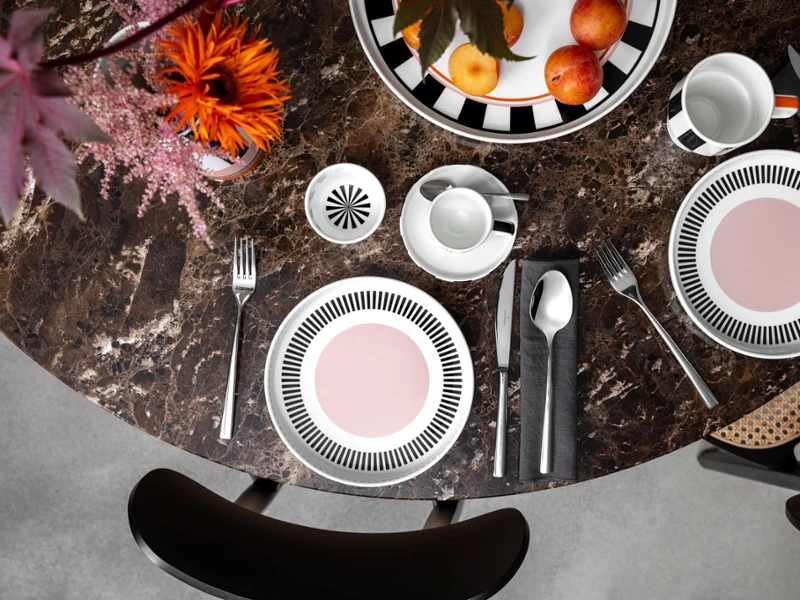 Colour explosion with Memphis by Villeroy & Boch A round marble table with two Memphis place settings, each with a plate, cutlery and a cup. A bowl of apples and a Memphis vase with flowers adorn the center.