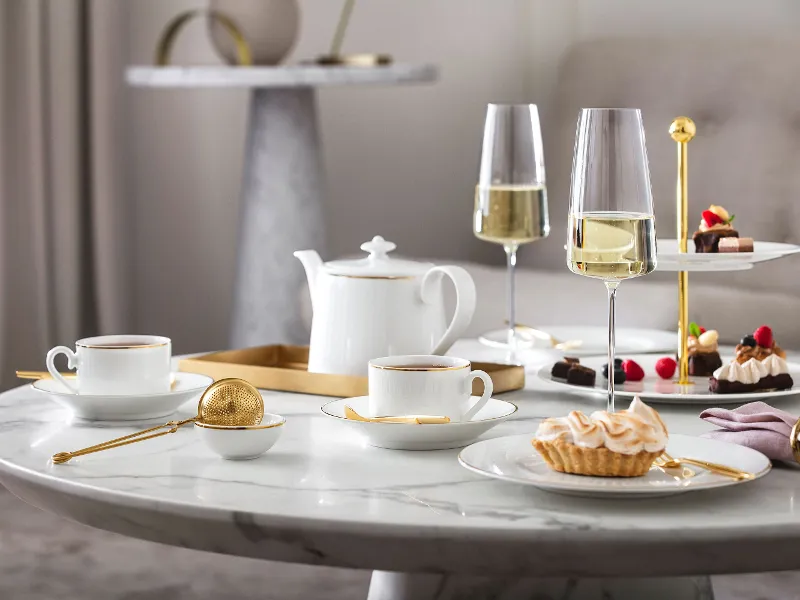 Une table en marbre avec des tasses à thé blanches Château Septfontaines, une théière, des flûtes à champagne MetroChic, des desserts et une passoire dorée.