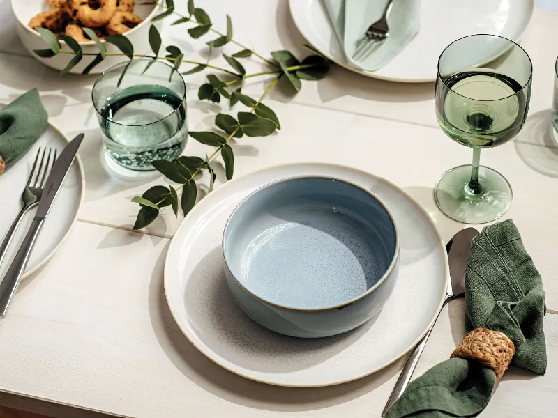 Een prachtig gedekte tafel met een lichtblauwe schaal van Villeroy & Boch Crafted op een wit bord, groene glazen, eucalyptustakken en groene stoffen servetten met ringen. Andere borden, bestek en voedsel zijn gedeeltelijk zichtbaar.