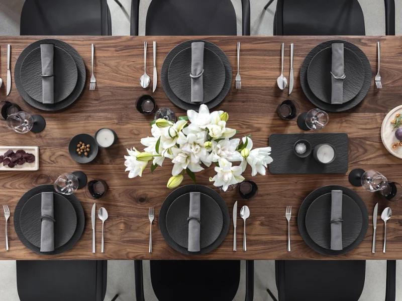A wooden dining table set for six with Manufacture plates, gray napkins, cutlery, and a white floral centerpiece.