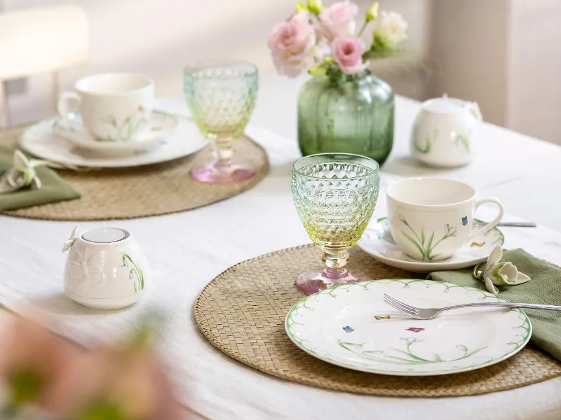Das Set Colourful Spring von Villeroy & Boch verschönert jeden Tisch für zwei mit Tellern und Tassen mit Blumenmuster, leuchtend grünen und gelben Gläsern, rosa Blumen in einer Vase und beigen Tischsets auf einer weißen Tischdecke.
