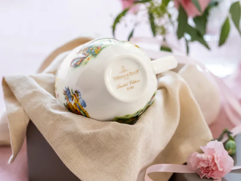 Een kopje uit de Annual Easter Edition van Villeroy & Boch ligt ondersteboven op een beige doek en toont de gouden jaarstempel, terwijl op de achtergrond een roze bloem en een vaas met bladeren te zien zijn.	
