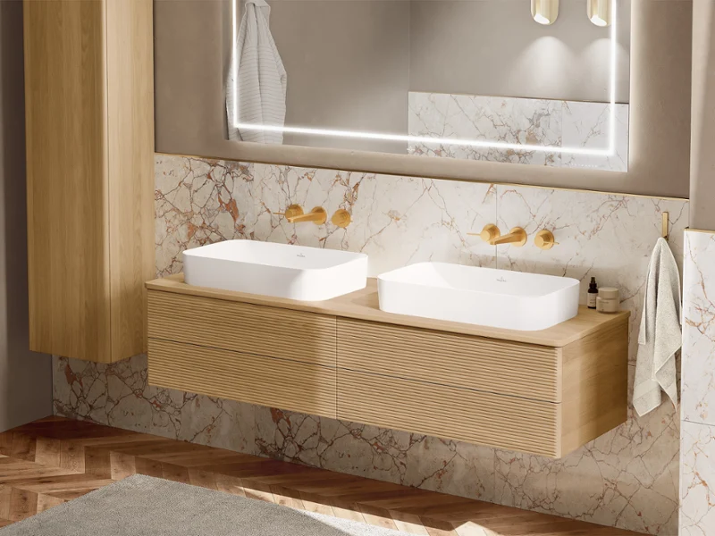 Two white countertop washbasins on a light wooden vanity unit. Behind it is a lighted mirror on the wall