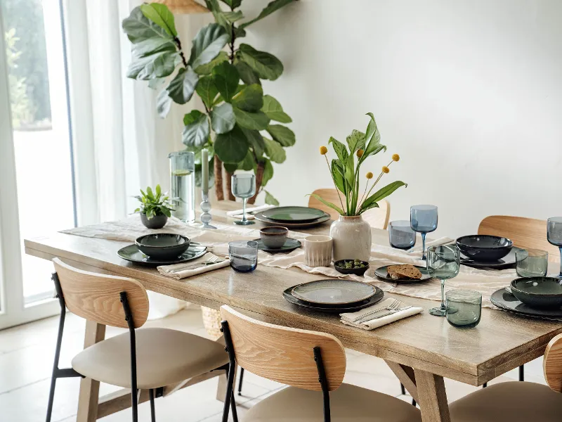 Dining table set for six with Lave collection plates by Villeroy & Boch, glasses, and plants as centerpiece in a bright room.
