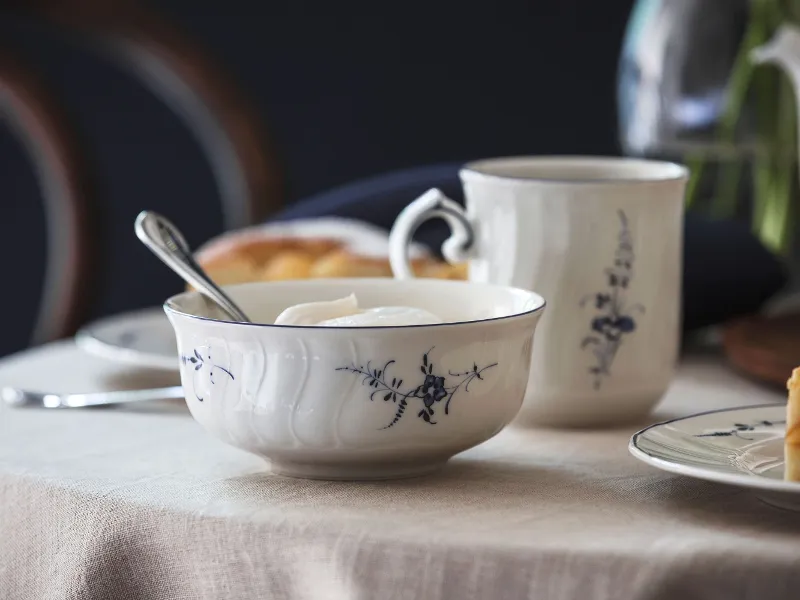 A table set with a Vieux Luxembourg bowl with yogurt and a spoon, a cup, and a plate on a beige tablecloth.