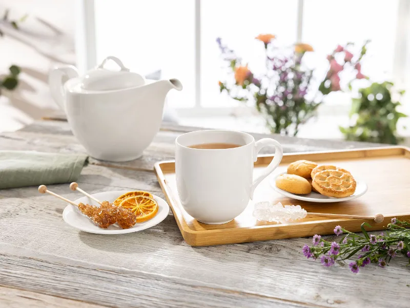 A Villeroy & Boch New Cottage teacup on a tray with cookies, a teapot, rock sugar, and dried orange slices on a wooden table adorned with flowers.