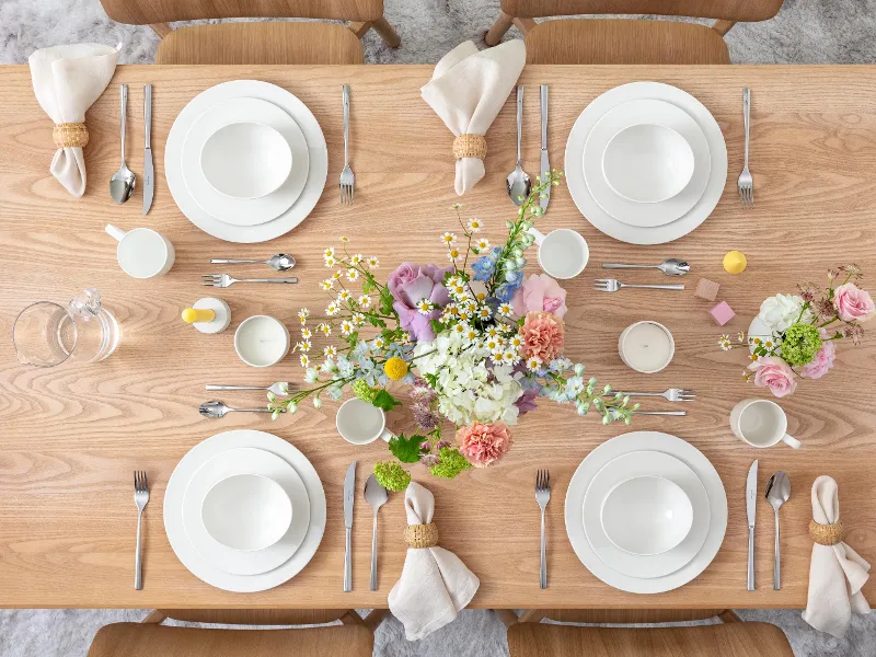 Une table à manger en bois dressée pour quatre personnes avec des assiettes blanches, des tasses et des couverts. Au centre, un arrangement floral avec des fleurs roses et blanches. Des carafes en verre sont prêtes, et des serviettes roulées sont placées à côté des assiettes.