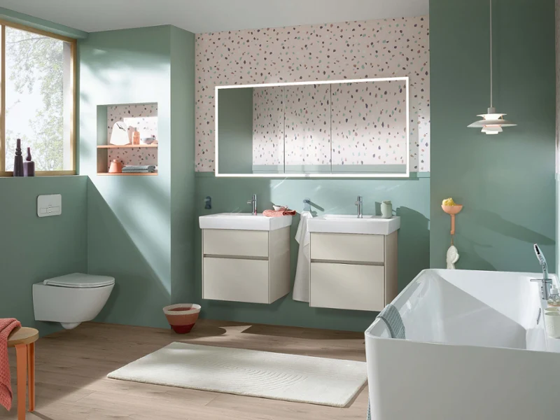 Salle de bain Collaro fraîche dans des tons pastel avec deux lavabos, une baignoire blanche et un mur style terrazzo. Des touches de vert doux et un sol en bois créent un look joyeux et moderne.
