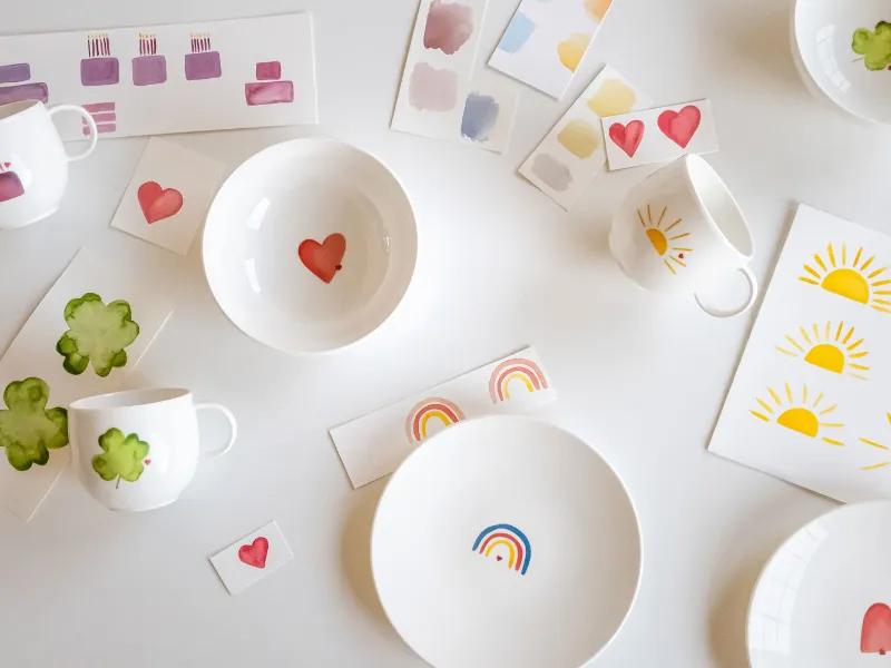 A set of Villeroy & Boch white "With Love" plates and cups, decorated with vibrant hearts, rainbows and tree patterns, elegantly arranged beside colour samples and pattern cards on a pristine white surface.