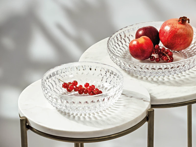 Two clear Boston glass bowls on marble tables with fruit. One contains pomegranates and an apple, the other contains red currants.